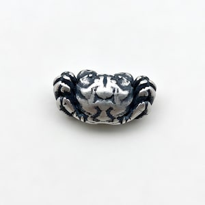 May include: A small, three-dimensional silver bead shaped like a crab. The metal has a polished finish on the raised surfaces and a dark, oxidized patina in the recessed areas to highlight the detailed texture of the crab's shell and legs.