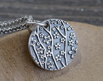 May include: A silver necklace with a round pendant featuring a detailed design of stylized trees and flowers.