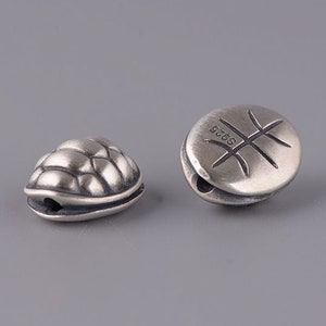 May include: Two small, silver-colored beads. One bead is shaped like a turtle shell, the other is round with a basketball design. Both beads have a small hole for stringing. The round bead has "S925" stamped on it.