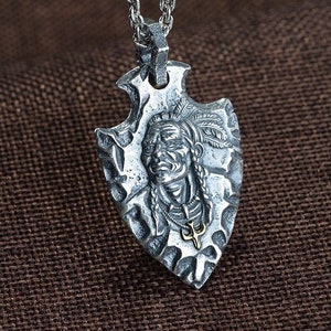 May include: Silver necklace with a pendant shaped like an arrowhead. The arrowhead features a detailed carving of a Native American chief's face.