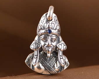 May include: Silver pendant featuring a detailed, ornate design of a mythical figure with a crown and a blue gemstone in the center of the forehead. The figure has a long, pointed tongue and a stern expression.