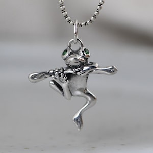 Può includere: Ciondolo a forma di rana argentata con occhi di pietra verde, che suona un flauto. La rana è sospesa a una collana a catena a sfera d'argento. Il design è dettagliato e fantasioso.