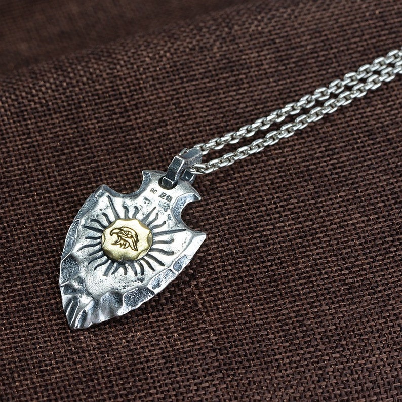 May include: Silver necklace with a silver arrowhead pendant featuring a gold eagle design. The arrowhead has a textured surface and the eagle is in the center of a sunburst design.