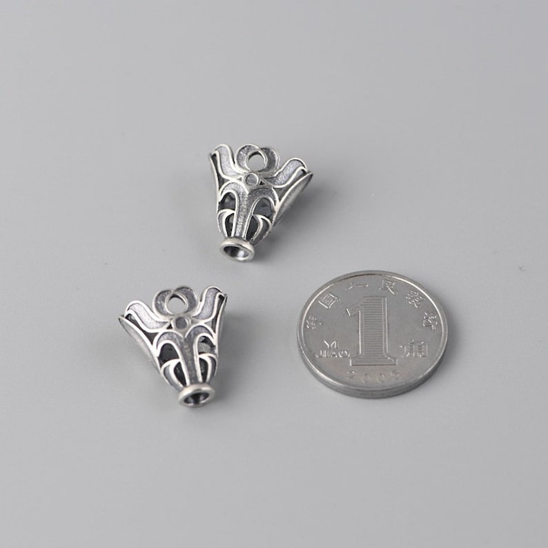 May include: Two silver-toned metal pendant caps with an intricate floral design. The caps are approximately the size of a Chinese yuan coin.