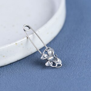 May include: A silver safety pin with a butterfly design. The pin is open and ready to be used.