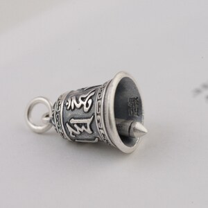 May include: A silver bell charm with intricate carvings and a small loop at the top. The bell has a pointed tip and is decorated with a repeating pattern around the rim.