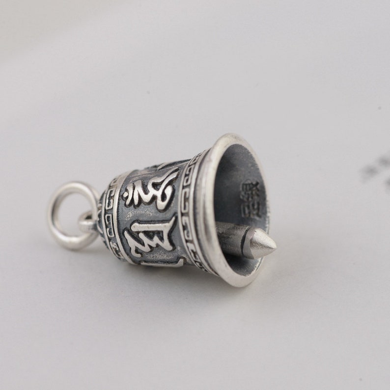 May include: A silver bell charm with intricate carvings and a small loop at the top. The bell has a pointed tip and is decorated with a repeating pattern around the rim.