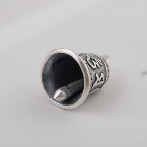 May include: Silver bell charm with a small silver rod inside. The bell is engraved with a series of symbols.