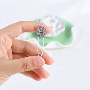 May include: A silver safety pin with a butterfly design. The pin is held in a hand.