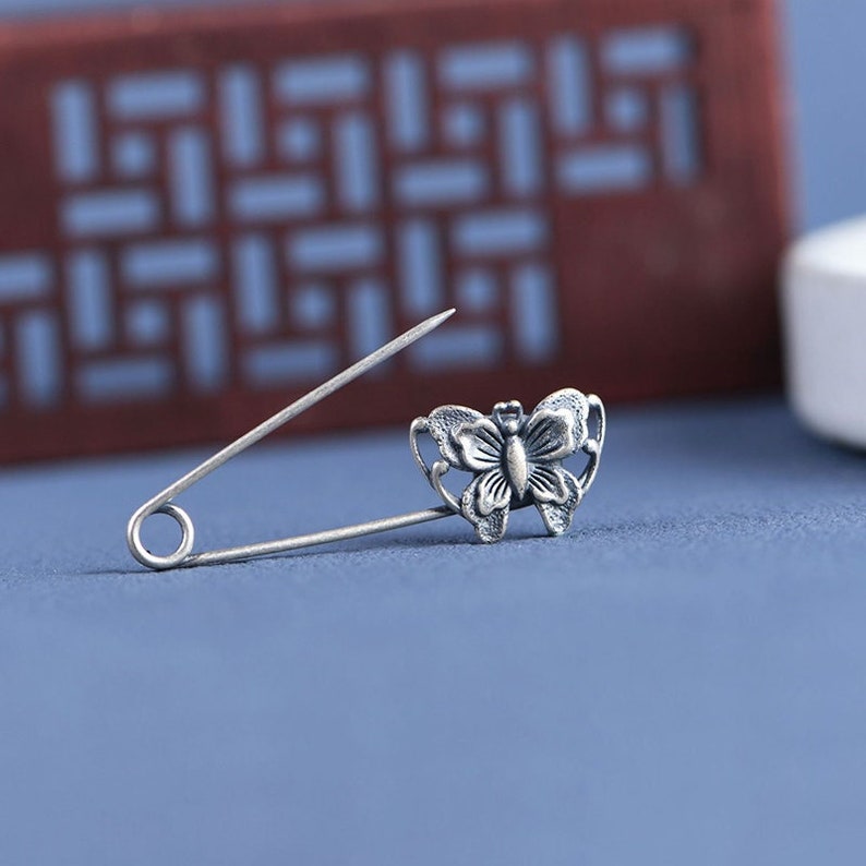 May include: A silver butterfly safety pin with a delicate design. The pin is open and ready to be used.