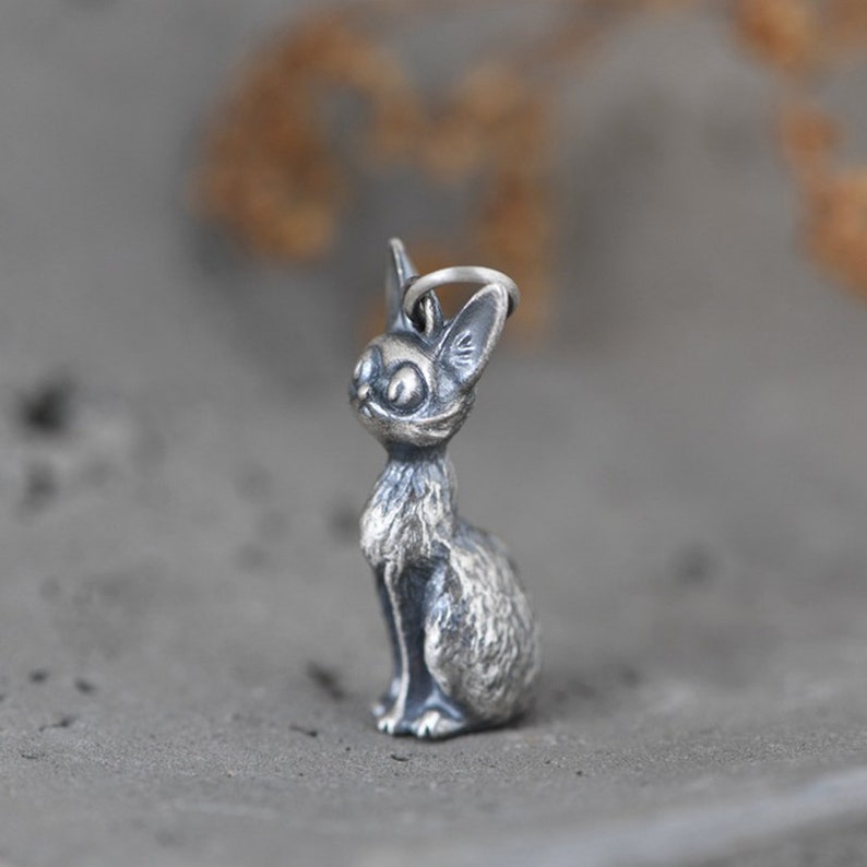 Puede incluir: Un amuleto de plata con forma de gato sentado. El gato tiene ojos grandes y un patr&oacute;n de pelaje texturizado y detallado.