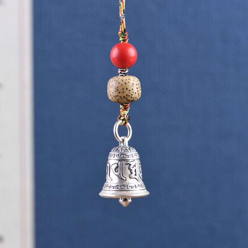 May include: Silver bell charm with Tibetan script, hanging from a red and brown cord with wooden and red beads.