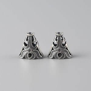 May include: Two silver-toned metal caps with intricate, ornate designs. The caps have a bell shape with a rounded top and a scalloped bottom.