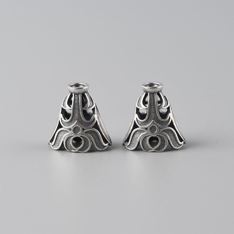 May include: Two silver-toned metal caps with intricate, ornate designs. The caps have a bell shape with a rounded top and a scalloped bottom.