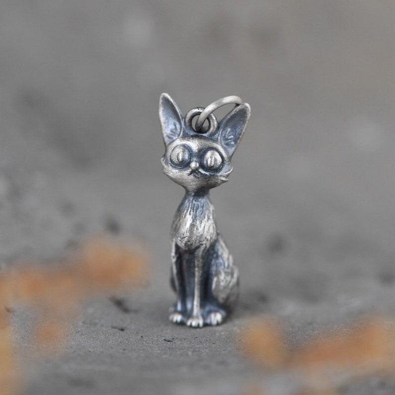 Puede incluir: Un amuleto de plata en forma de gato sentado con ojos grandes. El amuleto es detallado y tiene un aspecto realista.