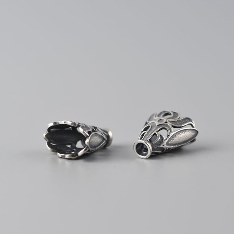 May include: Two silver-toned metal bead caps with an intricate, ornate design. The caps are open at one end and have a closed end with a small hole for threading.