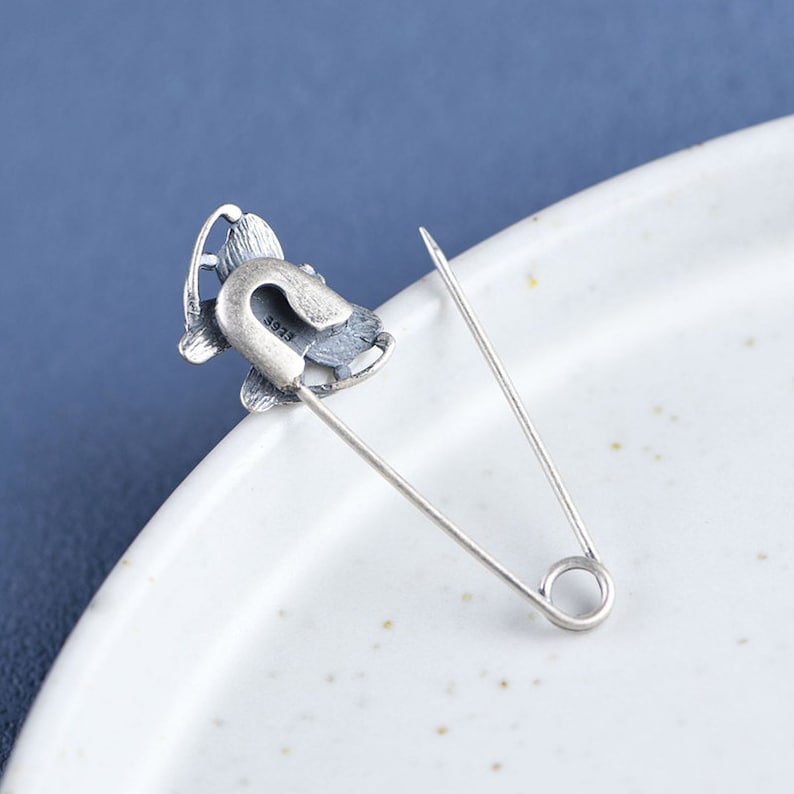 May include: Silver safety pin with a butterfly design. The butterfly has a textured surface and the pin has a silver finish. The back of the pin has the number "S925" engraved on it.