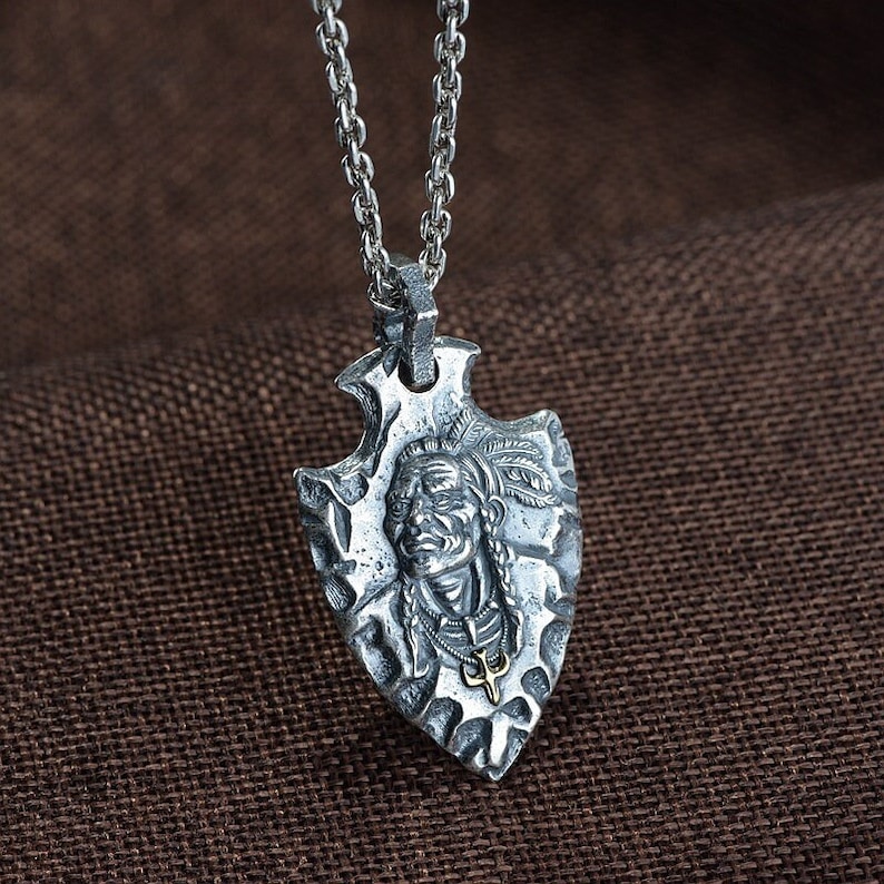 May include: Silver necklace with a pendant shaped like an arrowhead. The arrowhead features a detailed carving of a Native American chief's face.
