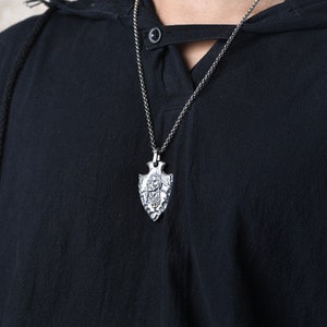 May include: A silver chain necklace with a silver arrowhead pendant. The arrowhead has a detailed design and a small stone in the center.