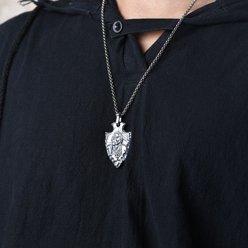 May include: A silver chain necklace with a silver arrowhead pendant. The arrowhead has a detailed design and a small stone in the center.