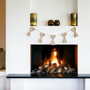 Op de afbeelding: Een witte open haard met een zwarte haard en een brandend vuur. Boven de open haard hangt een macramé slinger. Twee hoge, goudkleurige vazen en twee decoratieve glazen bollen staan op een plank.