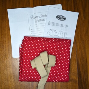May include: A sewing pattern for boxer shorts with a red and white polka dot fabric, a beige elastic band, and instructions for sizes XS to XL. The pattern is from DREDINI SEWING FACTORY SEATTLE.
