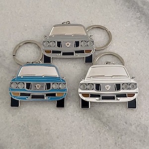 May include: Three metal keychains featuring a vintage car design. The keychains are silver, blue and white. Each keychain has a silver ring for attaching keys.