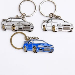 May include: Three car-shaped keychains in silver, white, and blue. Each keychain features a detailed car design with gold-colored wheels and a silver keyring. The keychains are displayed against a white background.