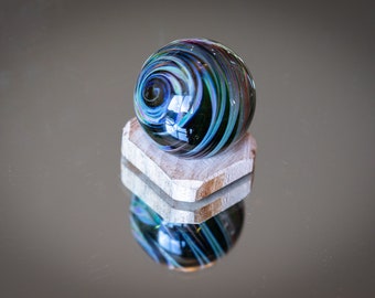 May include: A decorative glass marble with swirling blue, green, and red patterns, resting on a small, light-colored wooden stand. The marble's surface reflects light, creating a captivating visual effect. The stand is hexagonal in shape.