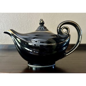 May include: A glossy black ceramic teapot featuring a gold-trimmed spout, a matching gold-accented lid with a pointed finial, and a distinctive curved handle that loops into a circular shape, creating a sleek and elegant silhouette for serving tea.