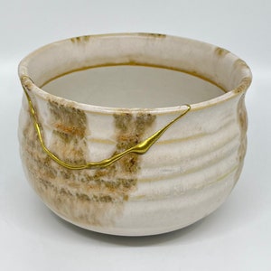 May include: A white ceramic bowl with brown and gold accents. The bowl has a rounded shape and a slightly flared rim. The gold accents are in the form of thin lines that run across the surface of the bowl.
