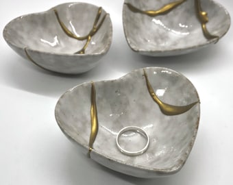 May include: Three white ceramic heart-shaped dishes with gold accents. The dishes are cracked and have a rustic look. The ring shown for scale is not included.