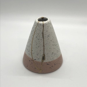 May include: A white and pink ceramic vase with gold accents. The vase has a speckled finish and a cone shape.