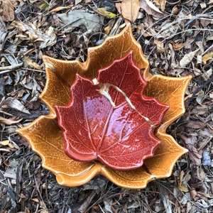 May include: Two ceramic leaf-shaped bowls, one inside the other. The outer bowl is a light brown color, and the inner bowl is a deep red color. Both bowls have a detailed leaf design.