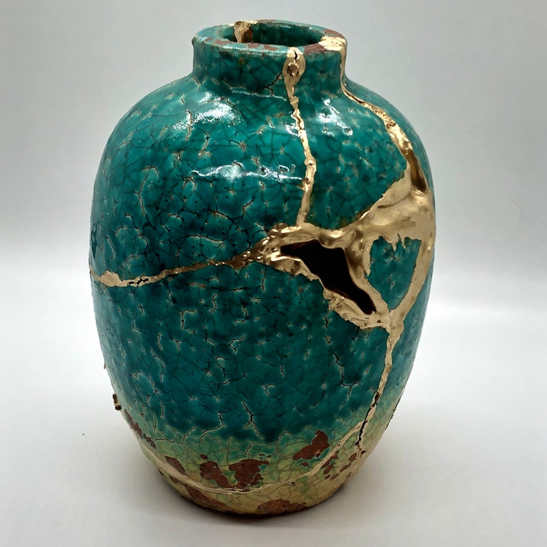 May include: A teal ceramic vase with gold crackle glaze and gold accents. The vase has a unique, distressed look.