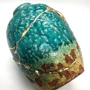 May include: A teal ceramic vase with a crackled glaze and gold accents. The vase has a rounded shape and a textured surface.