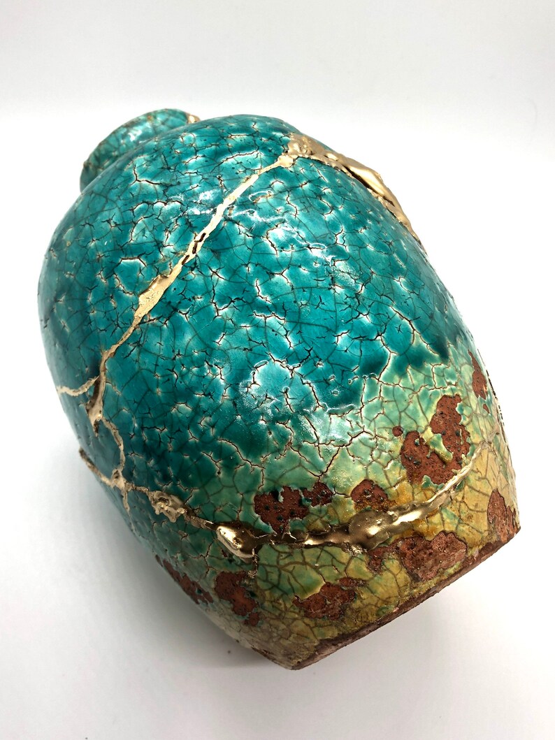 May include: A teal ceramic vase with a crackled glaze and gold accents. The vase has a rounded shape and a textured surface.