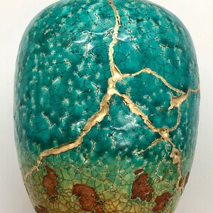 May include: A teal ceramic vase with gold cracks. The vase has a distressed look with a crackled glaze and gold accents.