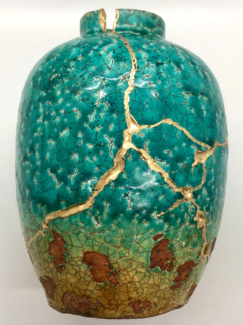 May include: A teal ceramic vase with gold cracks. The vase has a distressed look with a crackled glaze and gold accents.