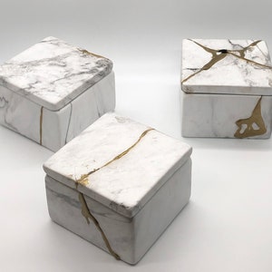 May include: Three square boxes with lids, each with a white marble-like finish and gold accents. The boxes are arranged in a triangular pattern on a white surface.