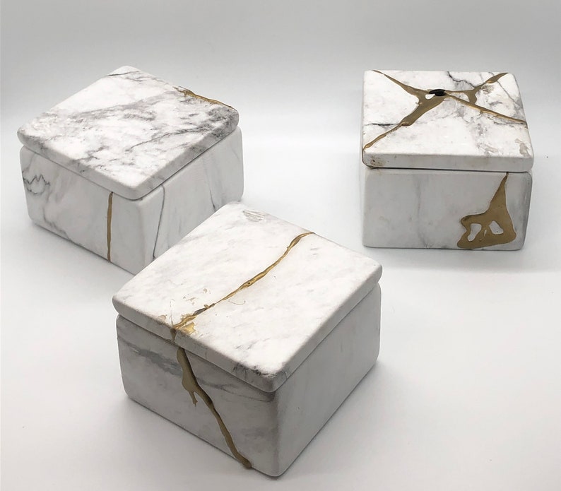 May include: Three square boxes with lids, each with a white marble-like finish and gold accents. The boxes are arranged in a triangular pattern on a white surface.