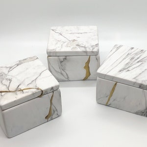 May include: Three white ceramic boxes with gold accents. The boxes have a marble pattern and are stacked on top of each other.