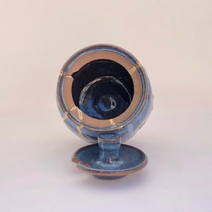 May include: A blue ceramic jar with a lid, featuring gold accents and a crackled glaze. The jar is open and the lid is sitting next to it.