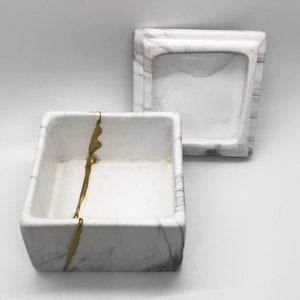 May include: White square box with a gold stripe design. The box has a matching lid. The box and lid are made of ceramic and have a marble-like finish.