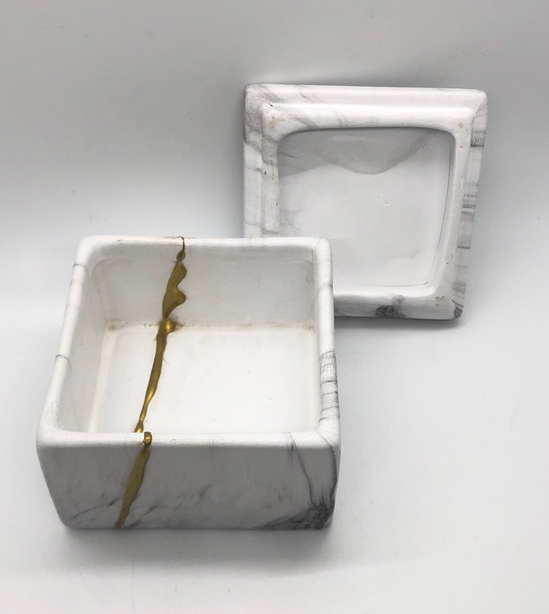 May include: White square box with a gold stripe design. The box has a matching lid. The box and lid are made of ceramic and have a marble-like finish.