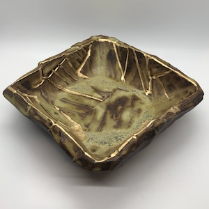 May include: A square ceramic bowl with a rough, textured exterior. The bowl has a muted green and brown color scheme, with gold accents. The interior of the bowl is a lighter green, and the gold accents create a striking contrast.