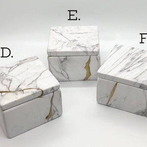 May include: Three white marble boxes with gold accents. The boxes are square and have lids. The boxes are labeled D, E, and F.