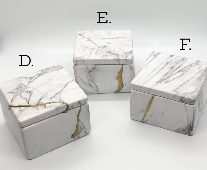 May include: Three white marble boxes with gold accents. The boxes are square and have lids. The boxes are labeled D, E, and F.