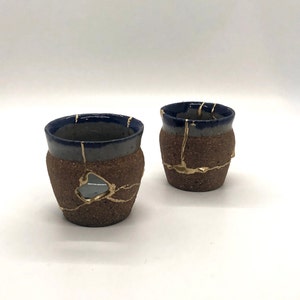 May include: Two small ceramic cups with a brown, speckled glaze and a blue rim. The cups have gold accents that look like cracks in the glaze.