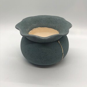 May include: A small, gray ceramic planter with a scalloped rim and a gold stripe. The planter has a textured surface and is perfect for small plants.